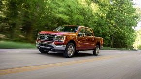 A metallic-red 2021 Ford F-150 Lariat four-door pickup truck traveling on a two-lane highway past thick trees on a sunny day