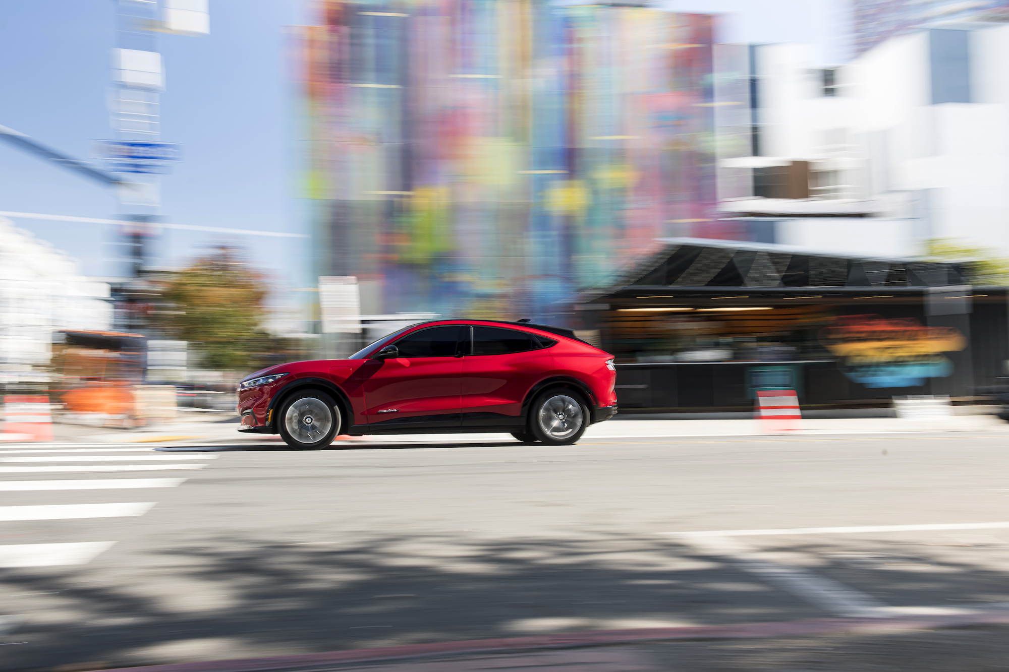 A red 2021 Ford Mustang Mach-E electric crossover SUV approaches a crosswalk at a city intersection on a sunny day