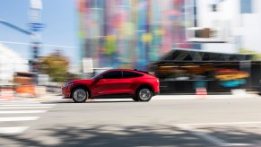 A red 2021 Ford Mustang Mach-E electric crossover SUV approaches a crosswalk at a city intersection on a sunny day