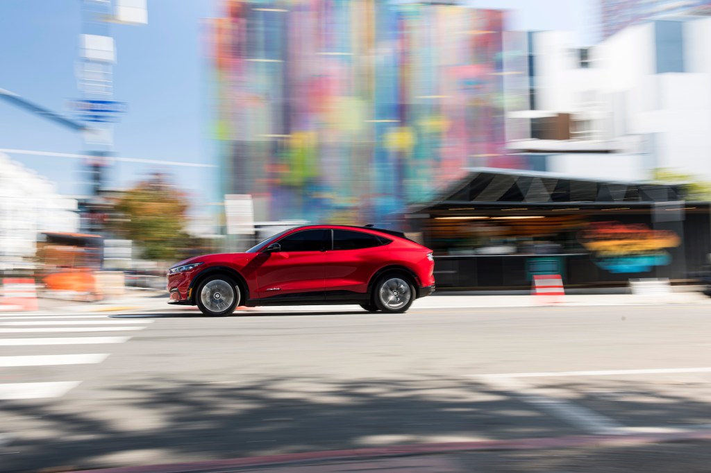A red 2021 Ford Mustang Mach-E electric crossover SUV approaches a crosswalk at a city intersection on a sunny day