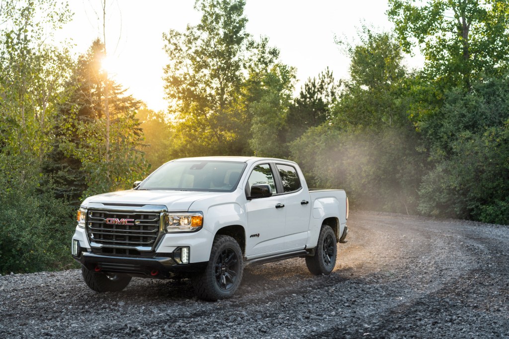 A white 2021 GMC Canyon AT4 Off-Road Performance Edition midsize pickup truck travels on a dusty gravel road through green trees and shrubbery