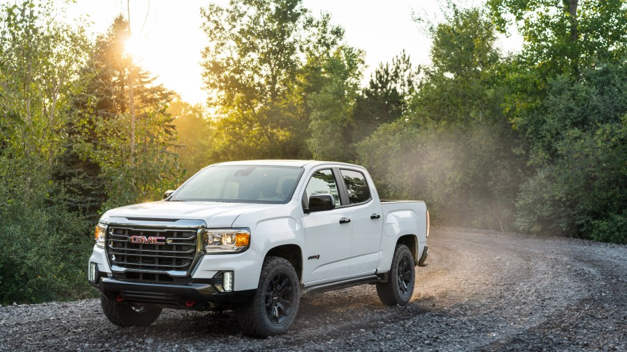 A white 2021 GMC Canyon AT4 Off-Road Performance Edition midsize pickup truck travels on a dusty gravel road through green trees and shrubbery