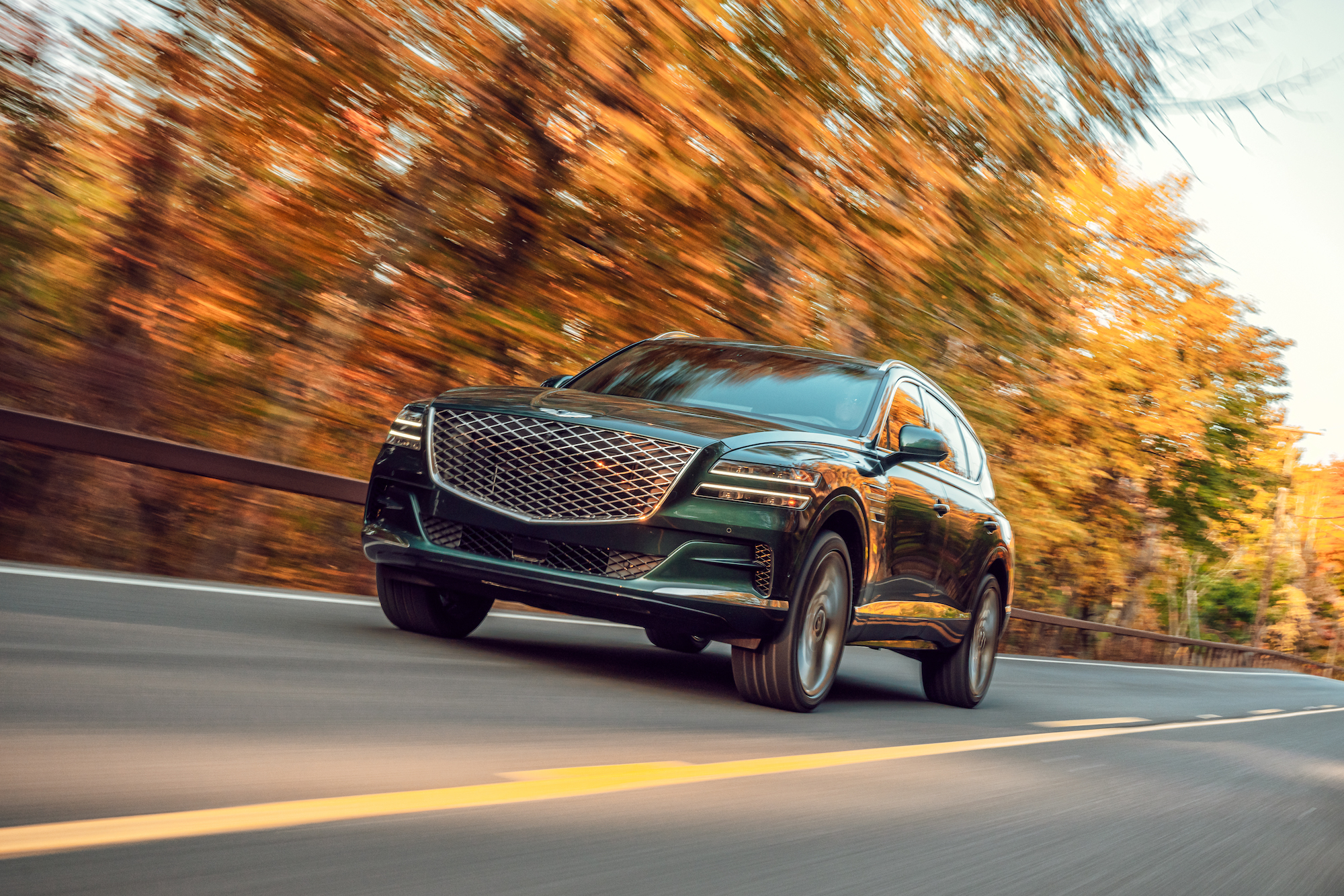 A dark-green 2021 Genesis GV80 travels on a tree-lined two-lane highway in Hudson Valley, New York