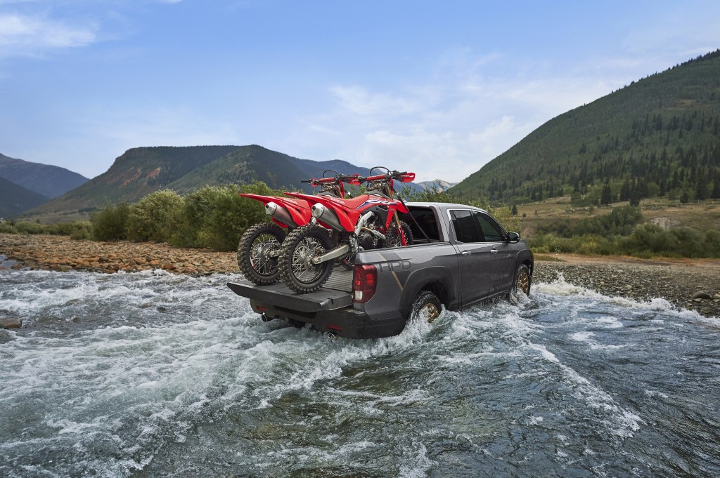 A black 2021 Honda Ridgeline HPD driving through shallow water with two red dirt bikes in the truck bed