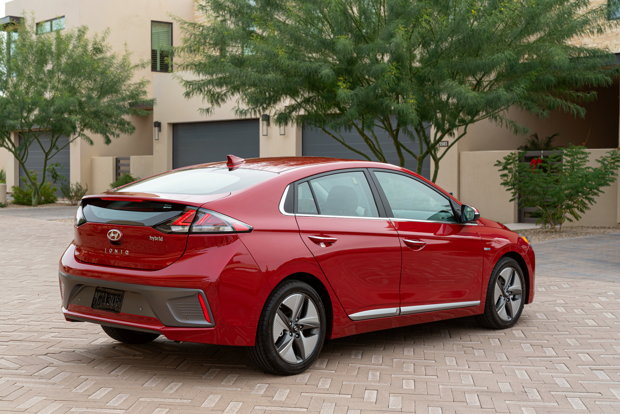 A red 2021 Hyundai Ioniq Hybrid four-door hatchback parked on beige brick pavers outside a tan-and-brown modern residence