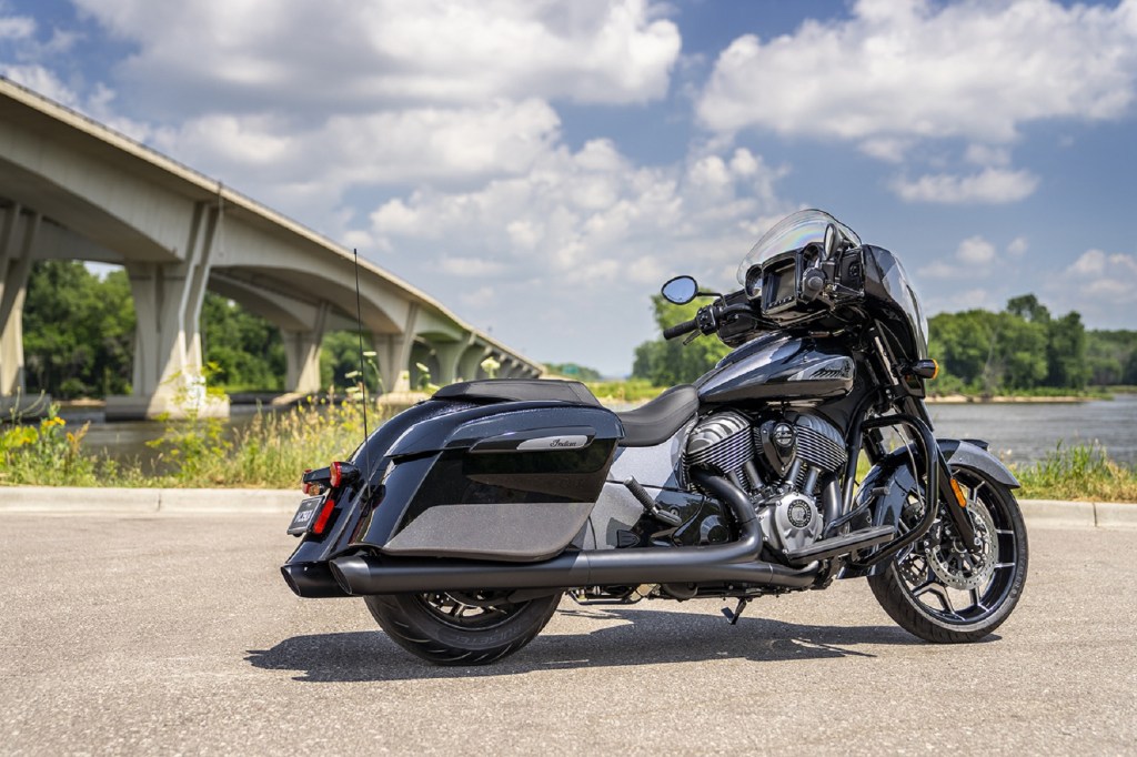 The rear 3/4 view of the metallic-black-and-gray 2021 Indian Chieftain Elite by a riverfront bridge