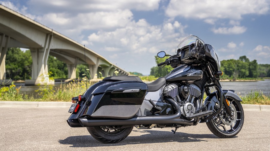 The rear 3/4 view of the metallic-black-and-gray 2021 Indian Chieftain Elite by a riverfront bridge