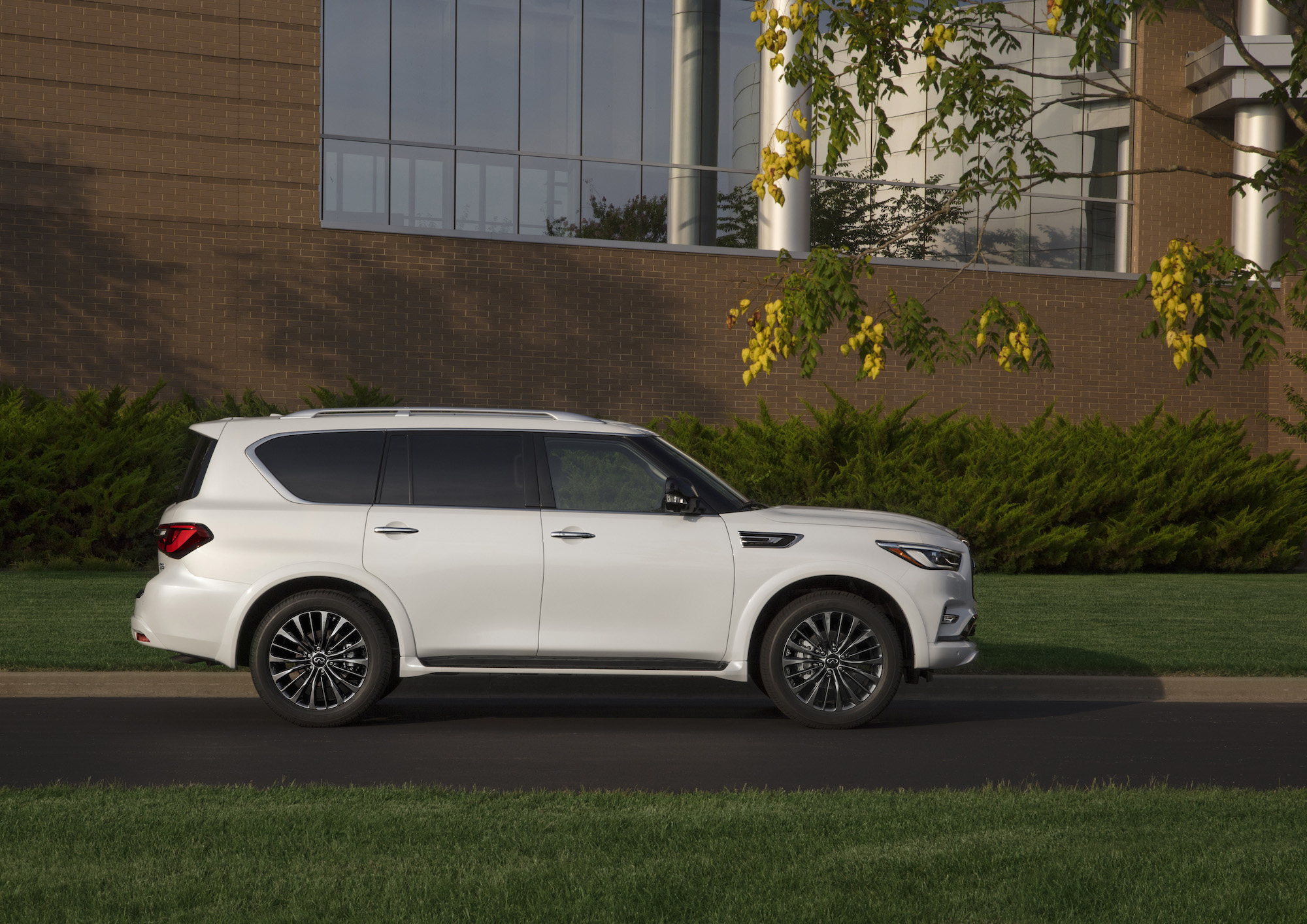 A white 2021 Infiniti QX80 three-row SUV parked along a curb next to grass and trees outside a modern office building