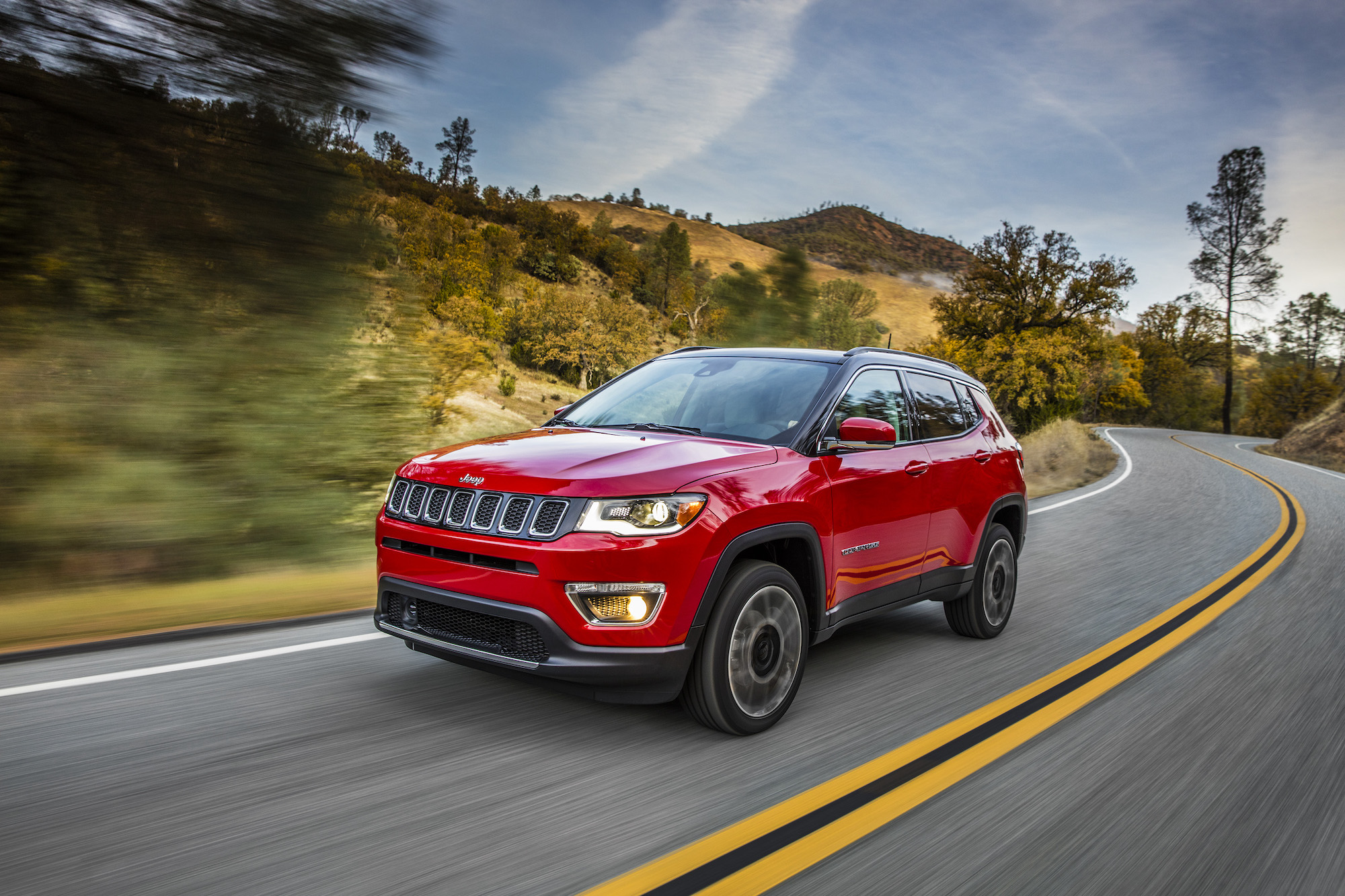 A red 2021 Jeep Compass Limited compact crossover SUV traveling on a two-lane highway through hills and trees