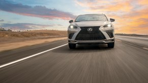 A silver 2021 Lexus NX300 compact luxury SUV travels on a two-lane highway with an expansive blue sky streaked with sun-kissed clouds in the background