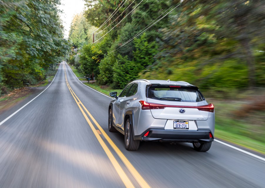A silver 2021 Lexus UX Hybrid luxury compact SUV travels on a tree-lined two-lane highway