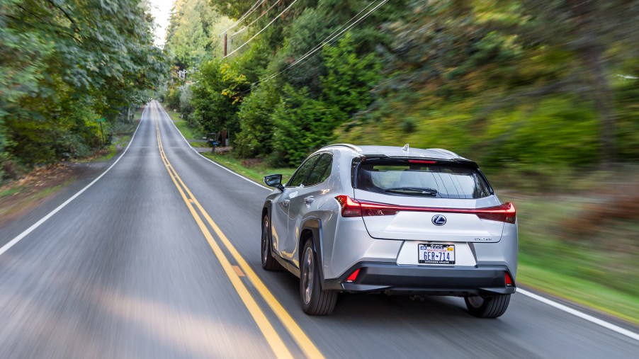 A silver 2021 Lexus UX Hybrid luxury compact SUV travels on a tree-lined two-lane highway