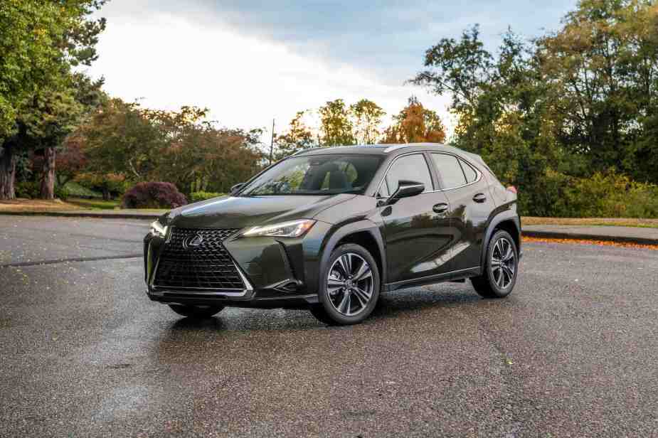 A 2021 Lexus UX with Nori Green exterior paint parked on a slab of asphalt in front of a sidewalk and trees with autumn leaves