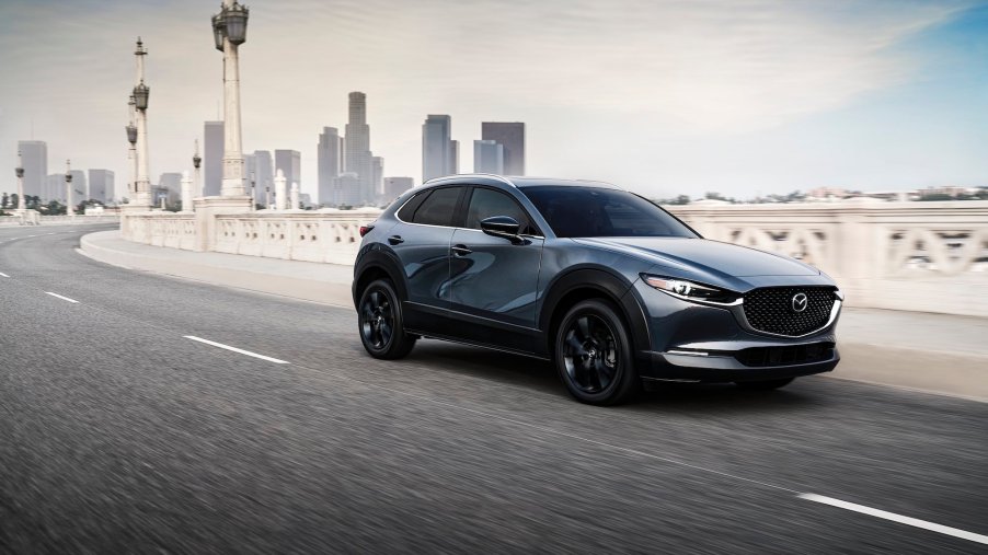 A metallic bluish-gray 2021 Mazda CX-30 2.5 Turbo subcompact crossover SUV travels on a bridge away from a city