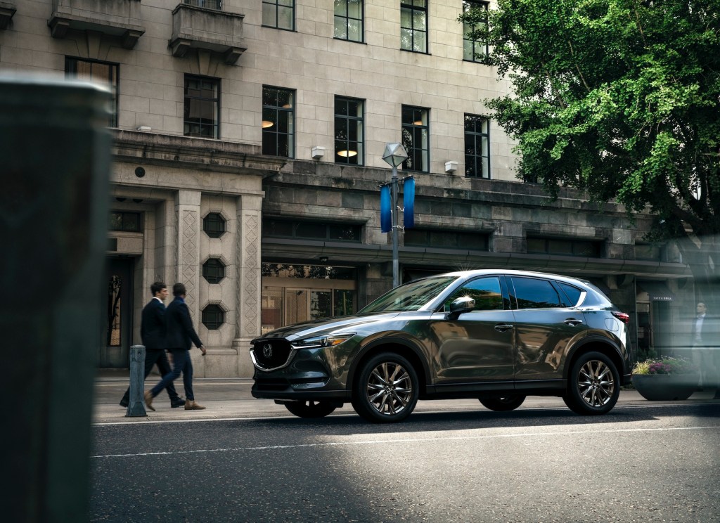 A dark-gray 2021 Mazda CX-5 compact crossover SUV parallel-parked outside a stone office building as two businesspeople walk by