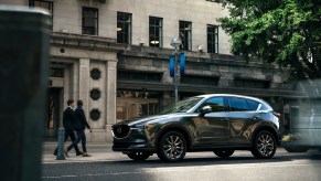 A dark-gray 2021 Mazda CX-5 compact crossover SUV parallel-parked outside a stone office building as two businesspeople walk by