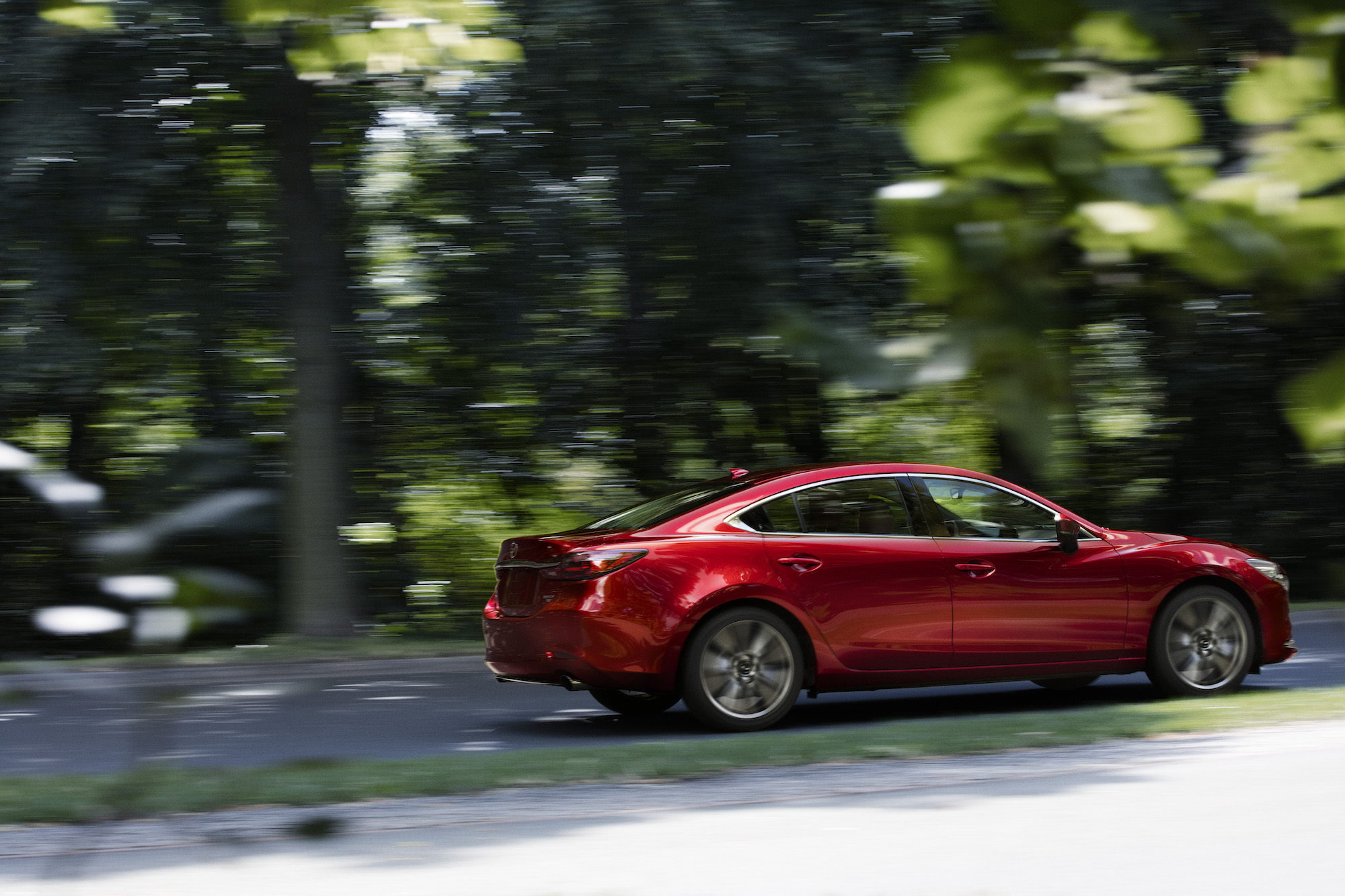 A red 2021 Mazda6 midsize sedan travels on a tree-lined road on a sunny day