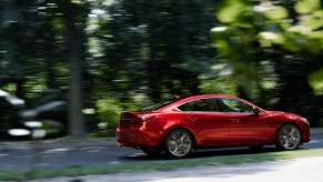 A red 2021 Mazda6 midsize sedan travels on a tree-lined road on a sunny day