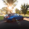 A blue 2021 Mercedes-AMG E63 S Wagon travels on a road lined with grass and trees on a sunny day