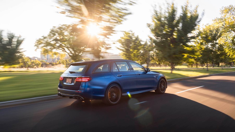A blue 2021 Mercedes-AMG E63 S Wagon travels on a road lined with grass and trees on a sunny day