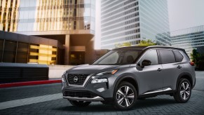 A dark-gray metallic 2021 Nissan Rogue compact crossover SUV parked outside glass skyscrapers