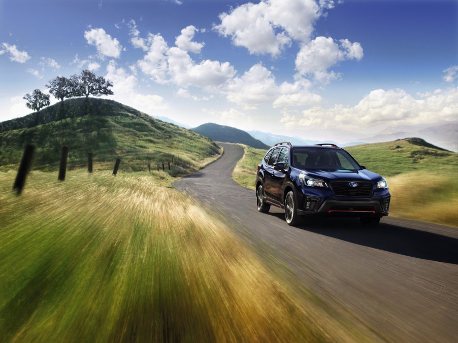A dark-blue 2021 Subaru Forester all-wheel-drive compact crossover SUV travels on a winding road through green rolling hills on a sunny day