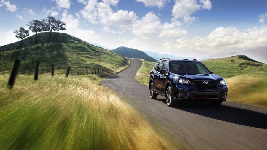 A dark-blue 2021 Subaru Forester all-wheel-drive compact crossover SUV travels on a winding road through green rolling hills on a sunny day