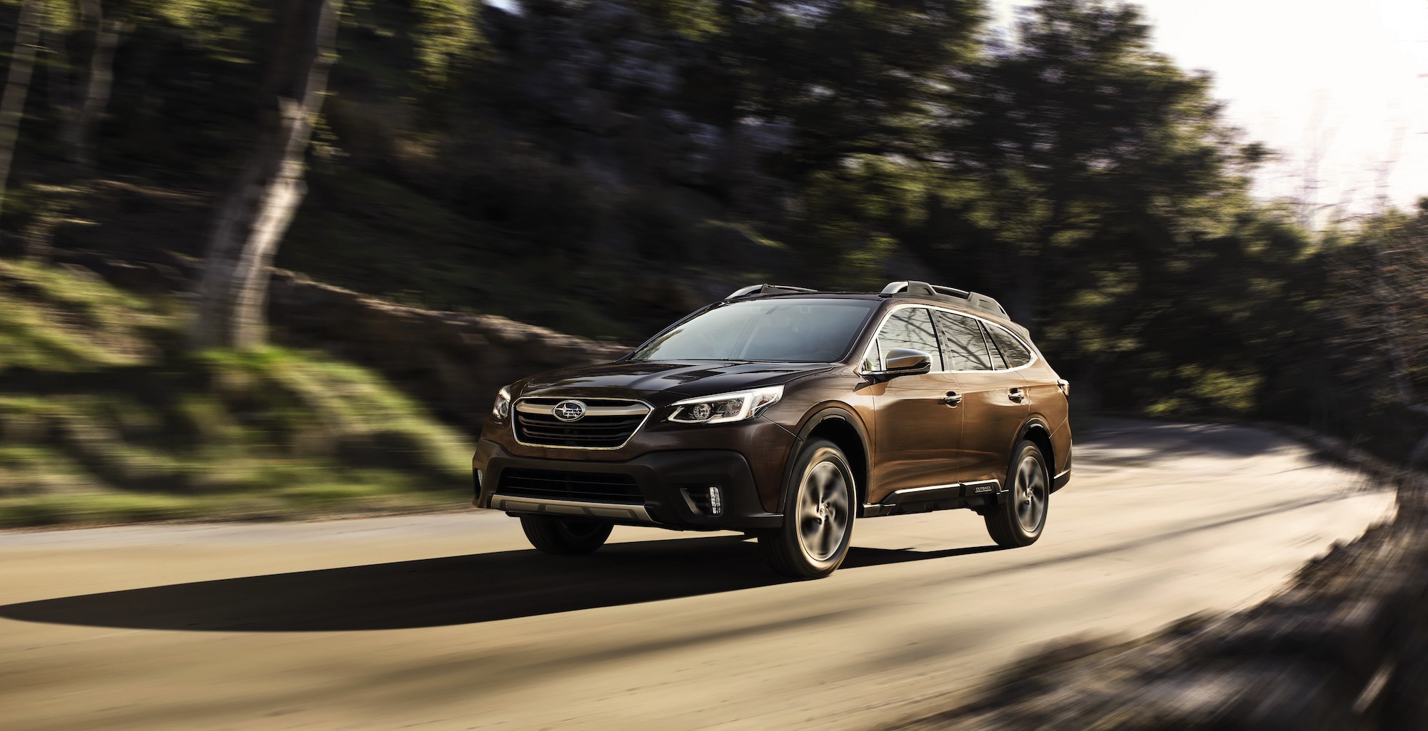 A brown 2021 Subaru Outback midsize two-row SUV traveling on a sun-dappled suburban road flanked by hills and trees