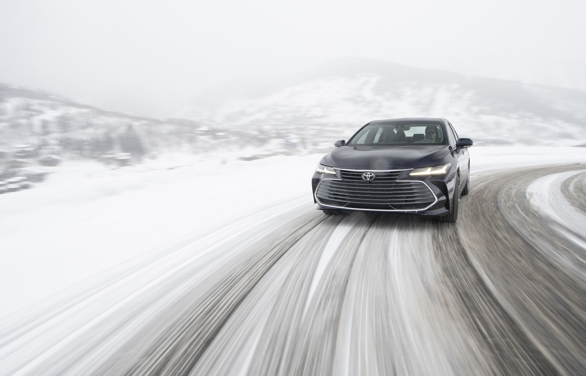 A dark-colored all-wheel-drive hybrid 2021 Toyota Avalon full-size sedan traveling on a snow-covered mountain road