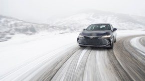 A dark-colored all-wheel-drive hybrid 2021 Toyota Avalon full-size sedan traveling on a snow-covered mountain road