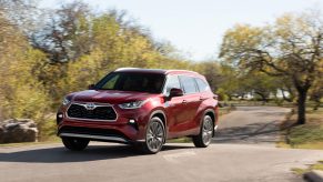 A metallic-red 2021 Toyota Highlander Hybrid midsize crossover SUV traveling on a road in a park with green grass and trees