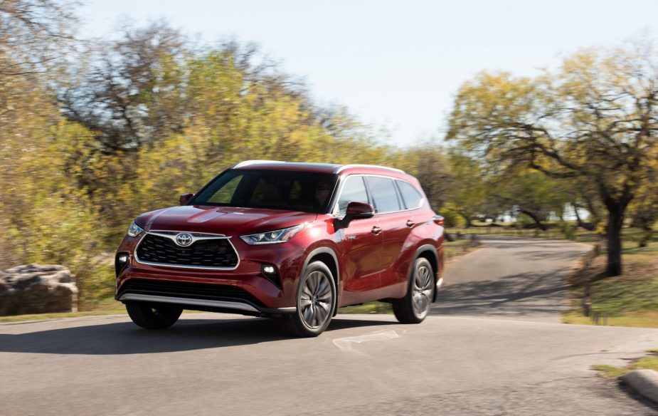 A metallic-red 2021 Toyota Highlander Hybrid midsize crossover SUV traveling on a road in a park with green grass and trees