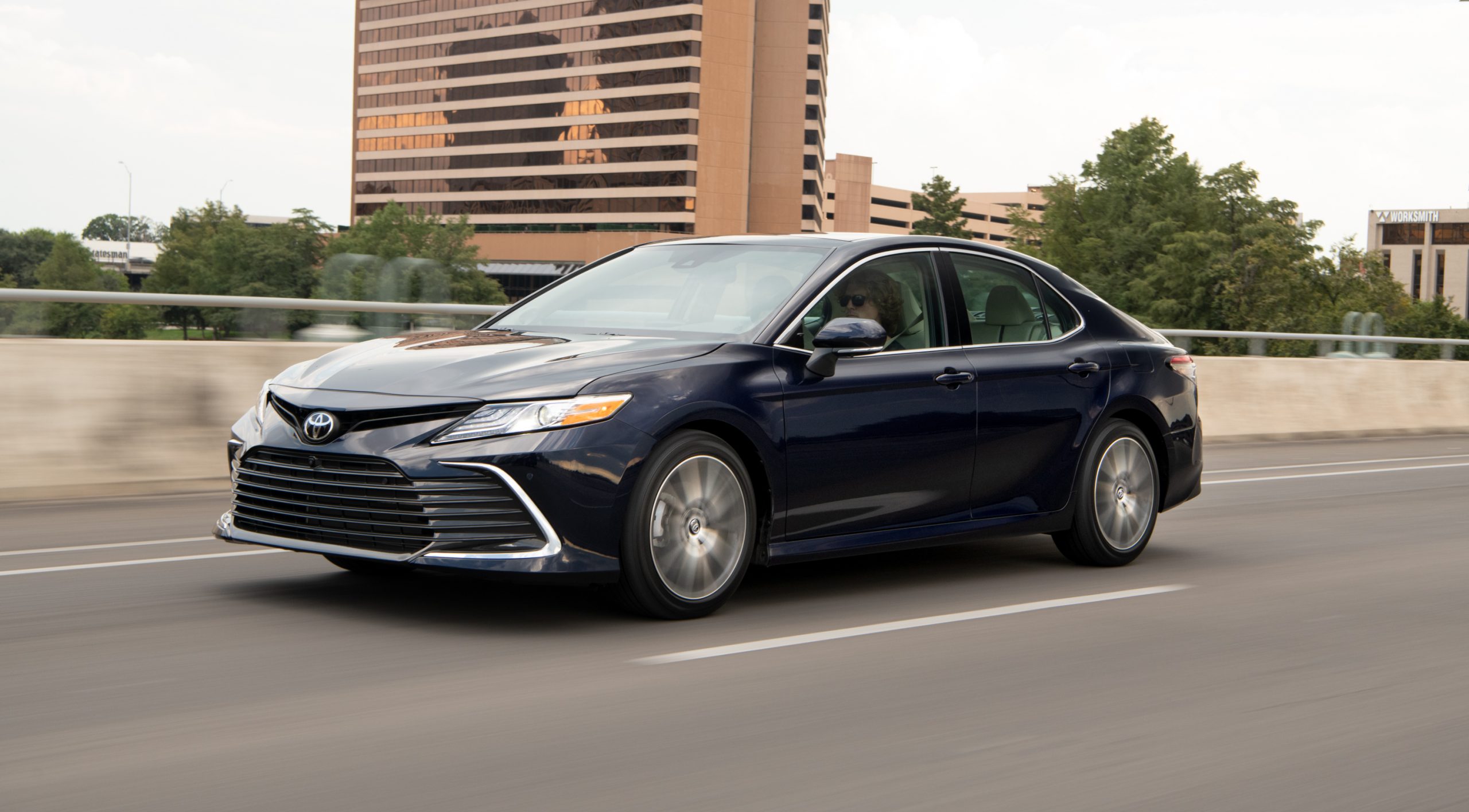 A blue 2021 Toyota Camry XLE driving down a highway road with a cityscape in the background