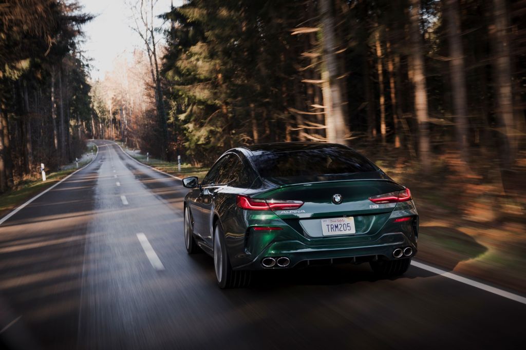 The rear view of a dark-green 2022 BMW Alpina B8 Gran Coupe driving down a road through a pine forest