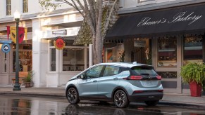 A light-blue metallic 2022 Chevrolet Bolt EV hatchback parked on a wet city street outside a bakery