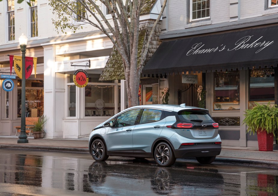 A light-blue metallic 2022 Chevrolet Bolt EV hatchback parked on a wet city street outside a bakery