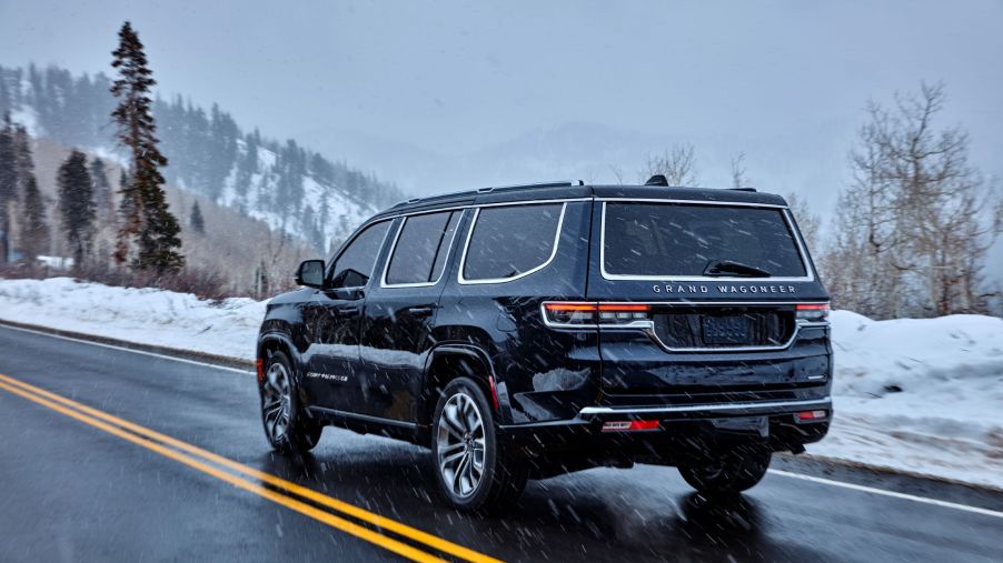 The rear 3/4 view of a dark-blue 2022 Jeep Grand Wagoneer Series III driving on a mountain road as snow falls