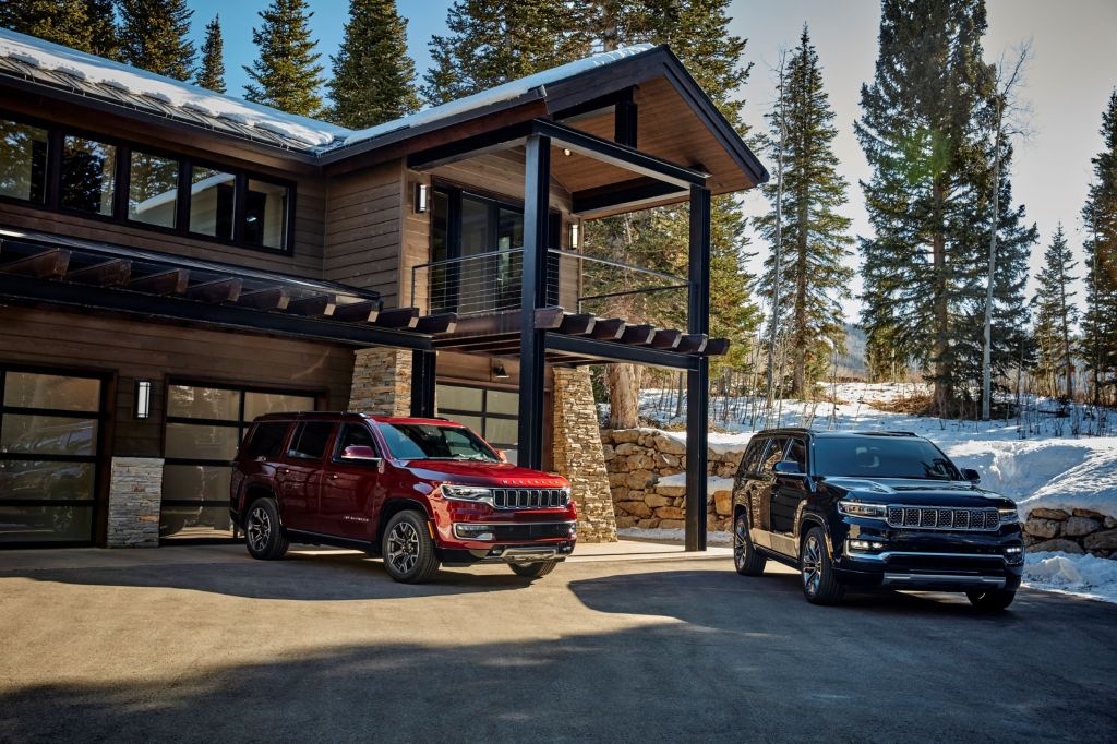 A red 2022 Jeep Wagoneer Series II next to a dark-blue 2022 Grand Wagoneer Series III by a wooden forest cabin