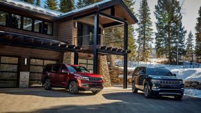 A red 2022 Jeep Wagoneer Series II next to a dark-blue 2022 Grand Wagoneer Series III by a wooden forest cabin