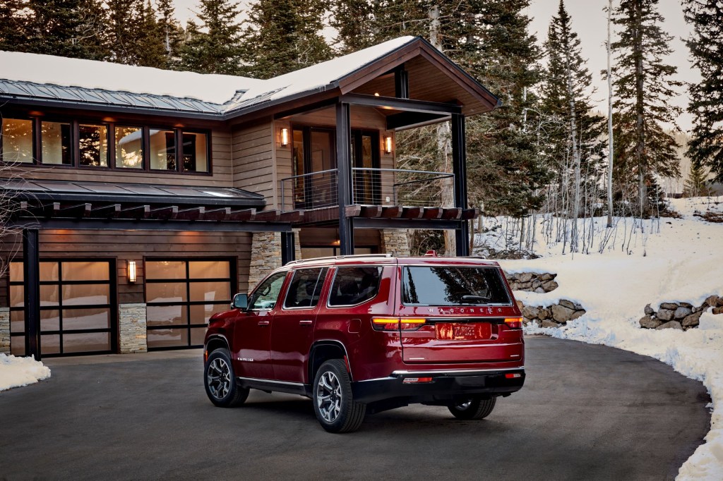 The rear 3/4 view of a red 2022 Jeep Wagoneer Series II next to a wooden cabin in a snowy forest