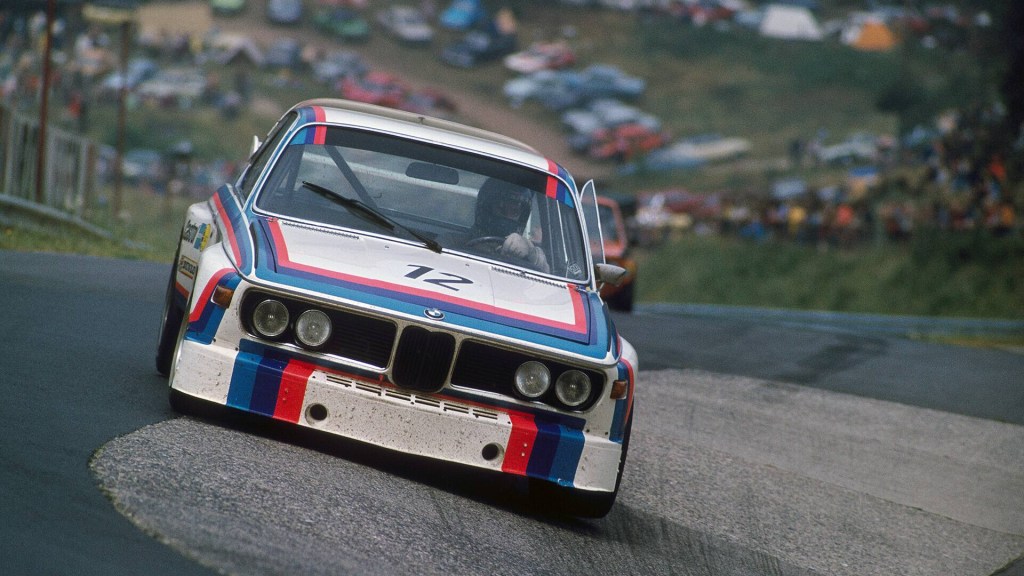 A white BMW 3.0 CSL 1973 with iconic red, purple and blue racing stripes taking a corner of a racetrack