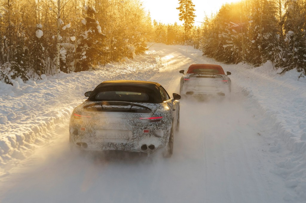 The rear view of a black-roofed and a red-roofed camouflaged 2022 Mercedes-AMG SL driving through a snow-covered forest