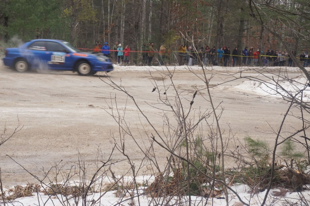 A blue Subaru Impreza WRX sliding at a 2018 SnoDrift Rally forest stage as spectators watch