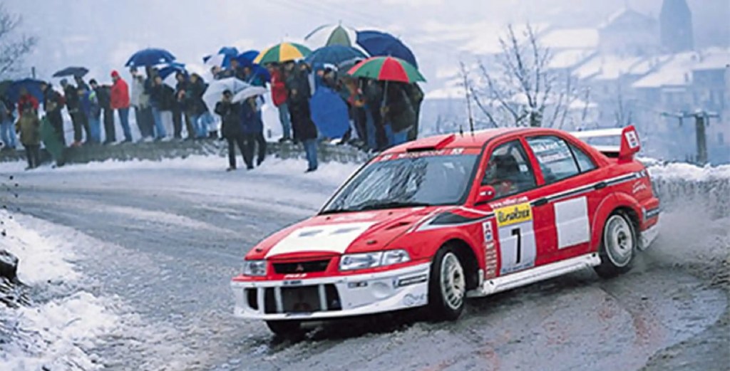 Tommi Makinen racing a red-and-white Mitsubishi Lancer Evo VI on a snowy rally stage with photographers behind him