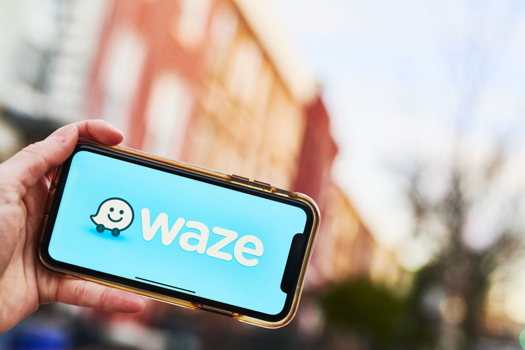 A hand grips a smartphone. The screen has a blue background and reads "WAZE" in large, white letters. The Waze logo, a smiling speech bubble on wheels, sits next to the name of the app