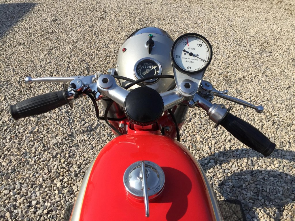 The handlebars, fuel tank, and dials of a red 1965 Ducati Mach 1 on a gravel driveway