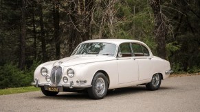 A white 1965 Jaguar S-Type 3.8 parked next to a forest