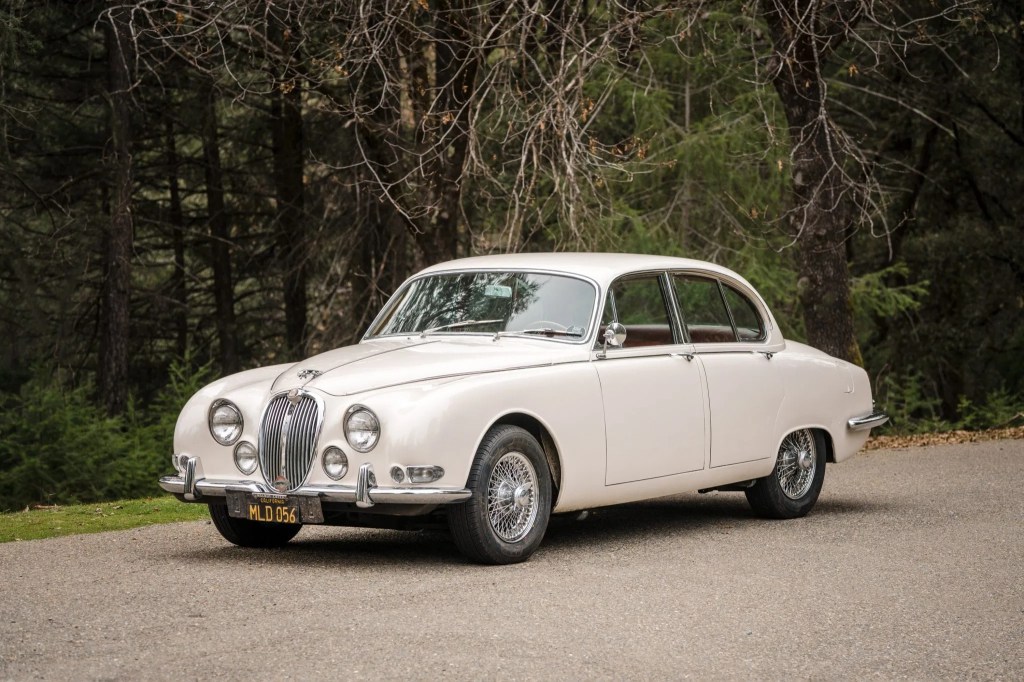 A white 1965 Jaguar S-Type 3.8 parked next to a forest