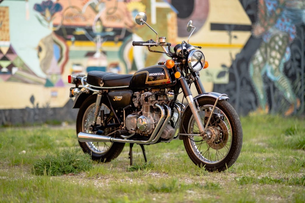 A black-and-gold 1973 Honda CB350F with an X-taped headlight in a yard next to a graffitied wall