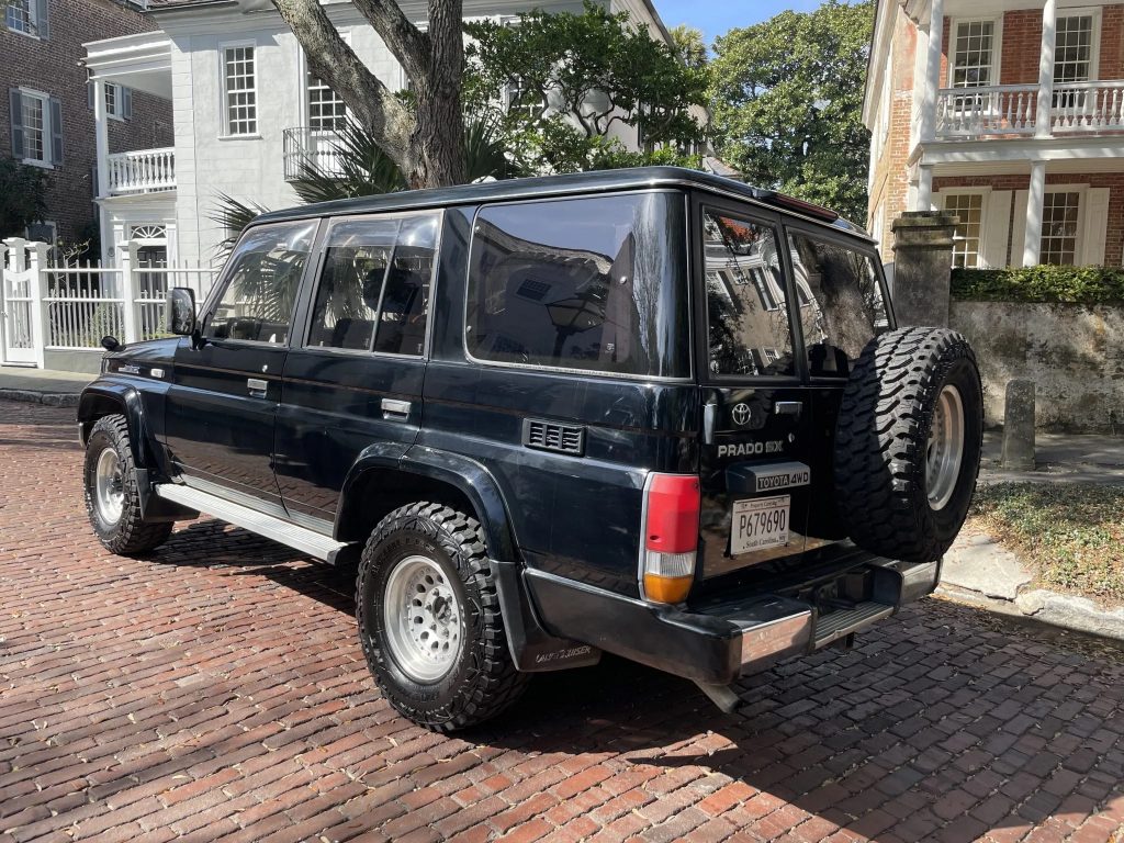 The rear 3/4 view of a black 1993 Toyota Land Cruiser Prado in front of some colonial-style houses